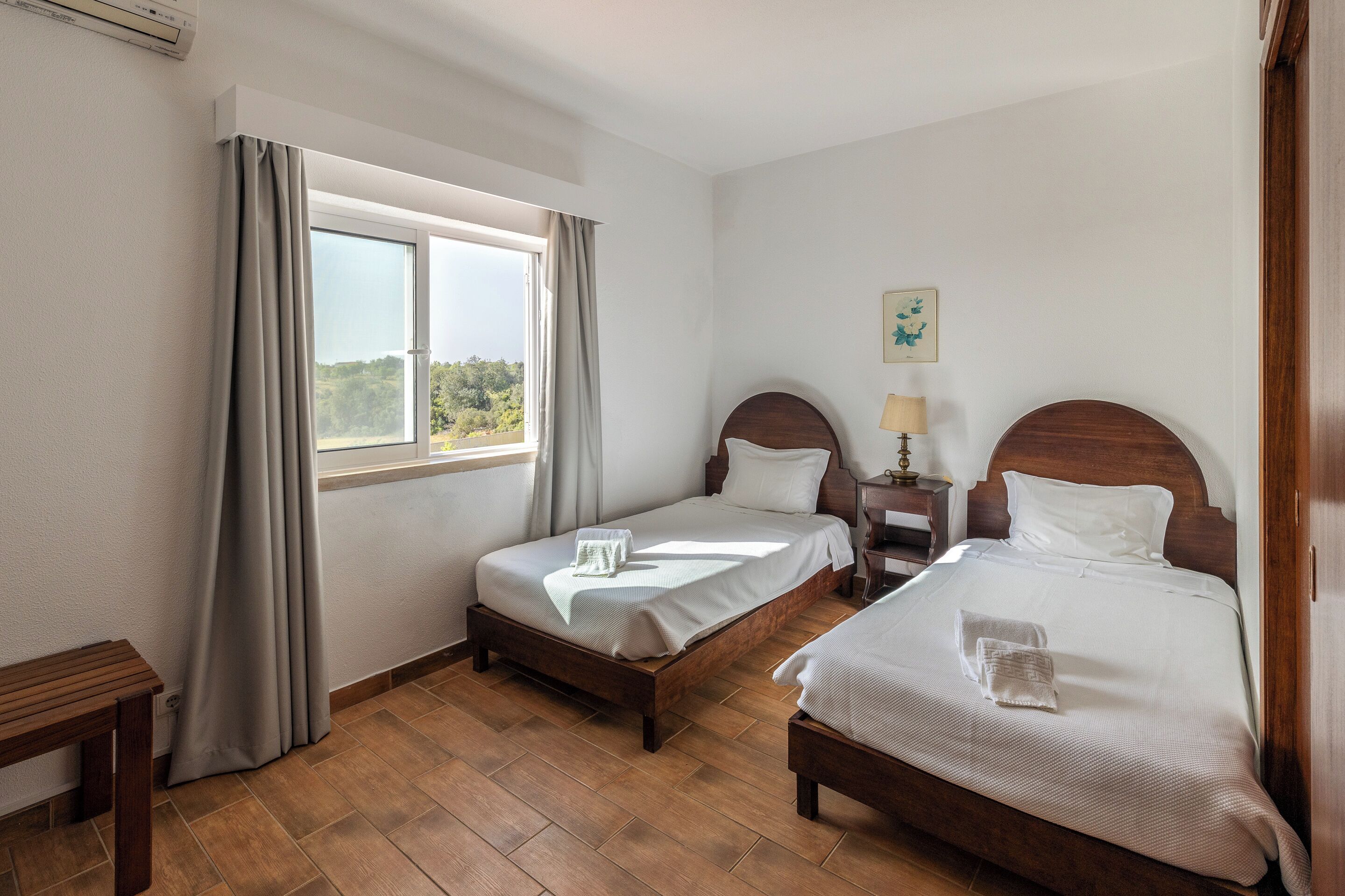 A hotel room with two beds, a wooden floor, a window with curtains, a bedside table, and a lamp.