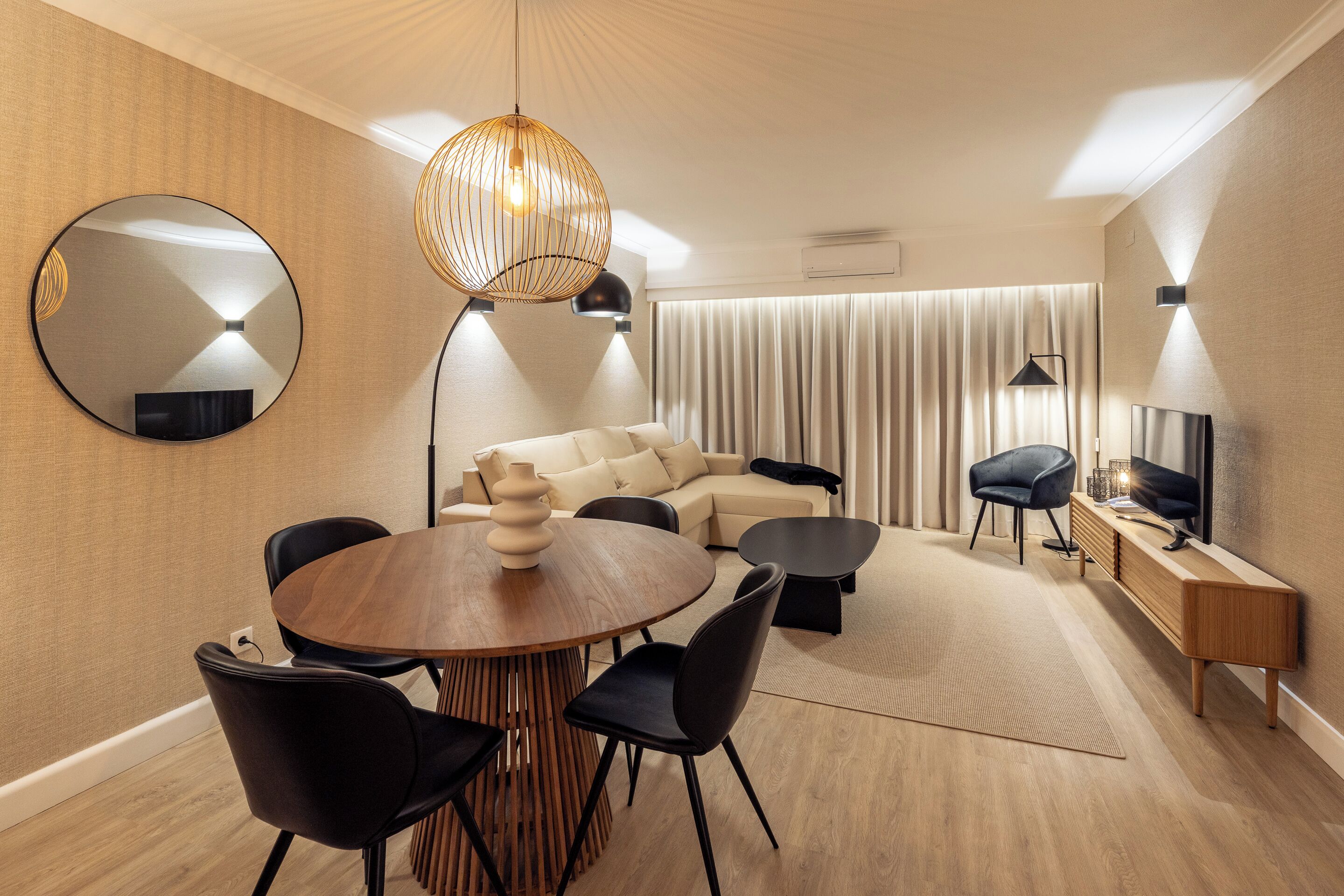 Una habitación de hotel moderna con una mesa de comedor redonda, sillas negras, un sofá, un televisor y un espejo grande.