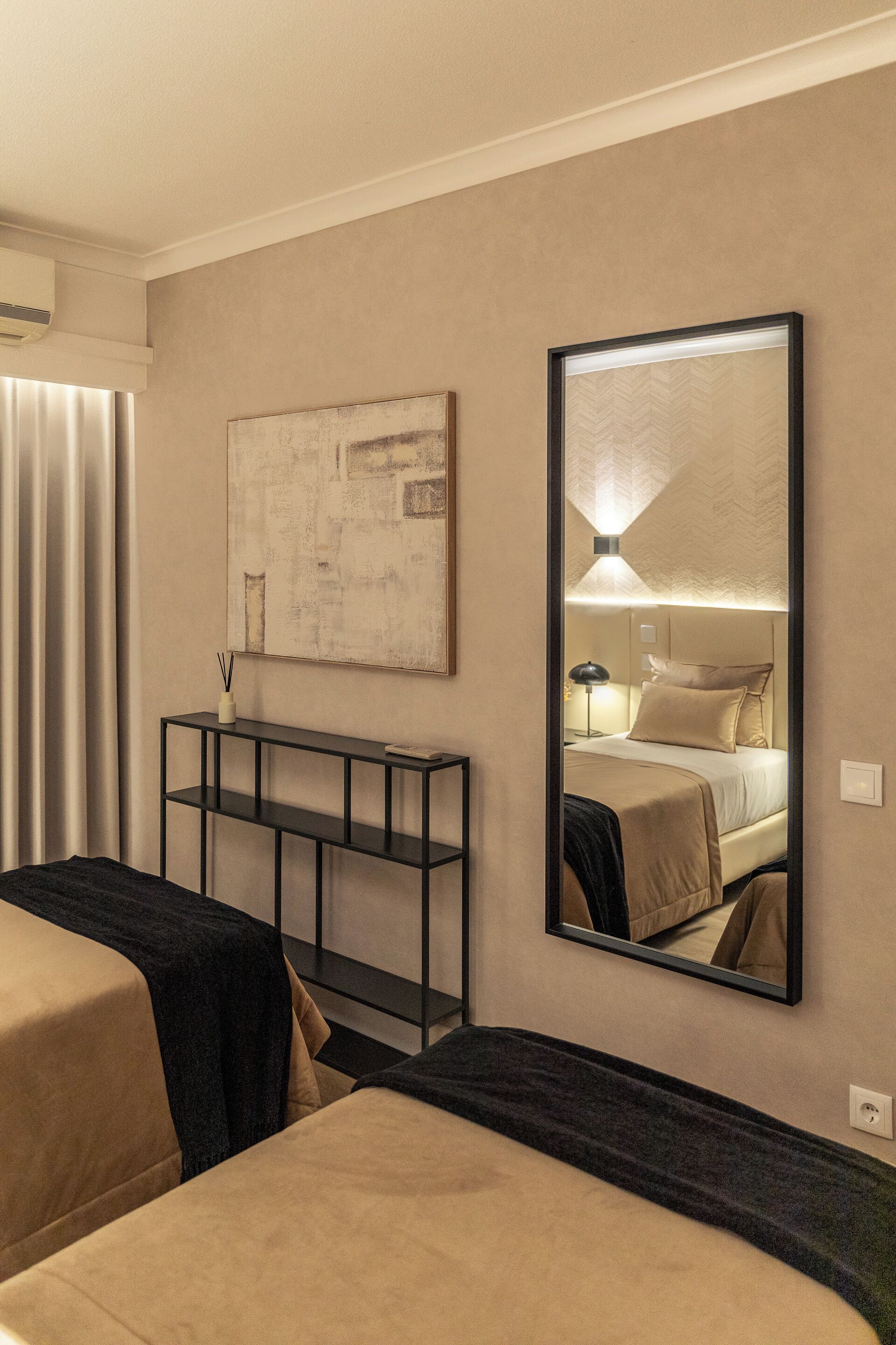 Ein Hotelzimmer mit einem großen Spiegel, der das Bett und ein an der Wand befestigtes Kunstwerk widerspiegelt. Ein Nachttisch mit einer Lampe und einer kleinen Vase mit Kerze sind ebenfalls zu sehen.
