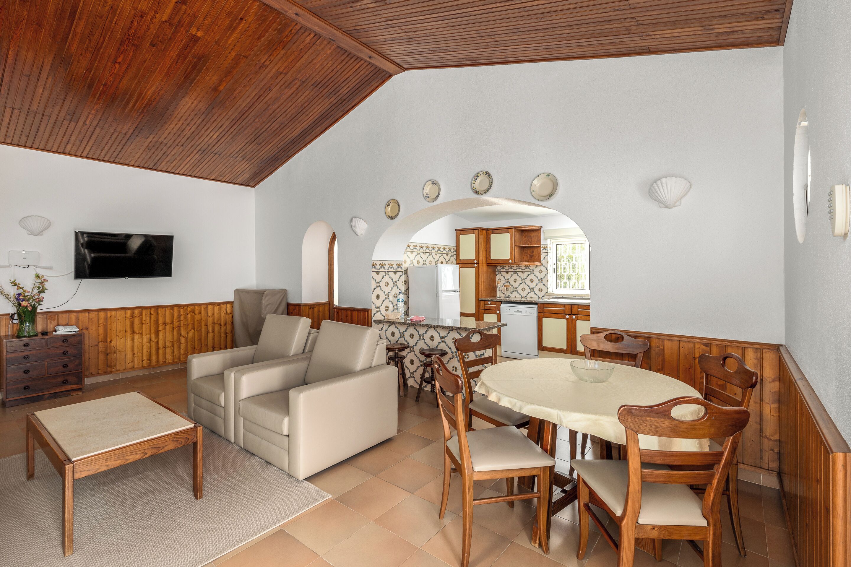 A spacious living area with a wooden ceiling, a sofa, a dining table with chairs, and a kitchen in the background.