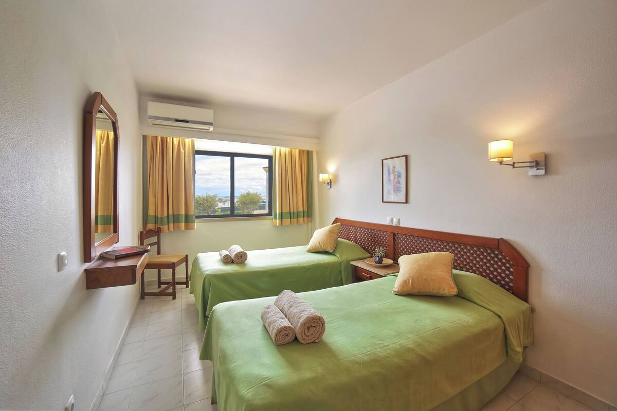 Habitación de hotel con dos camas, un escritorio, una silla, un ventanal con cortinas y una lámpara de pared.