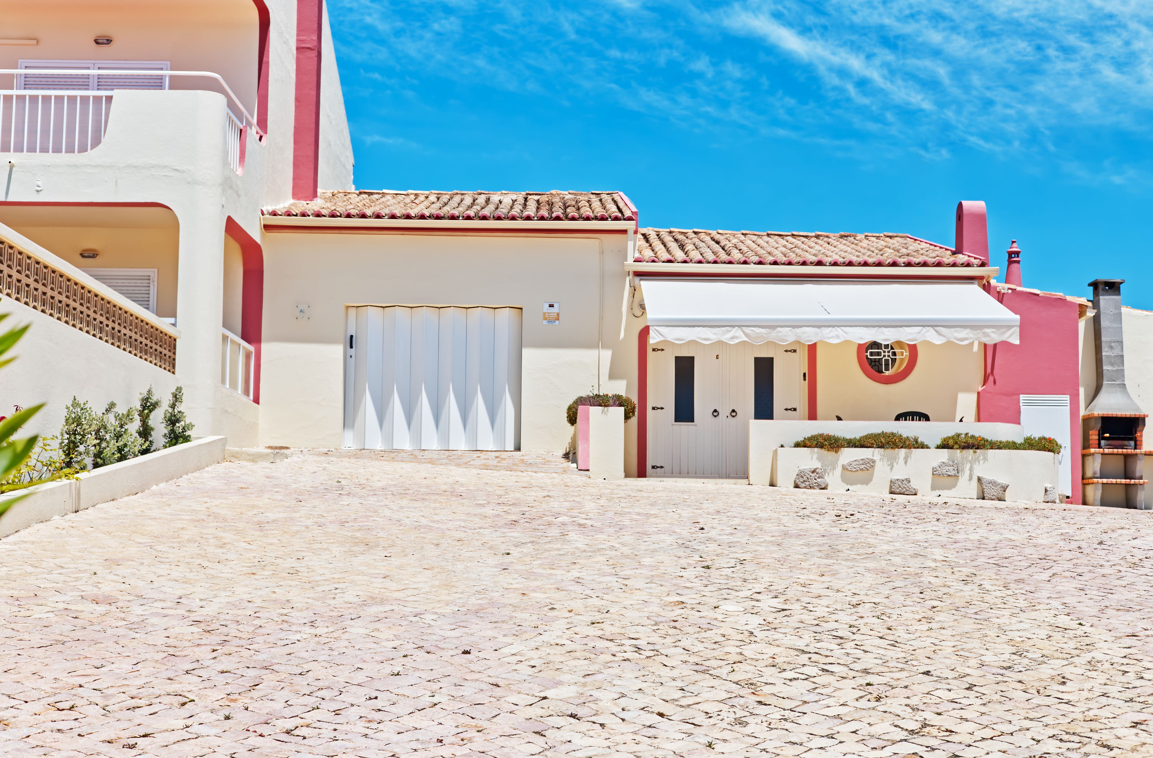 Una calle adoquinada que lleva a un edificio con fachada blanca y detalles rojos, un área de estar al aire libre techada y una chimenea.
