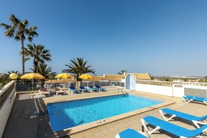 A rooftop pool with blue lounge chairs and yellow umbrellas, surrounded by palm trees and a clear sky.