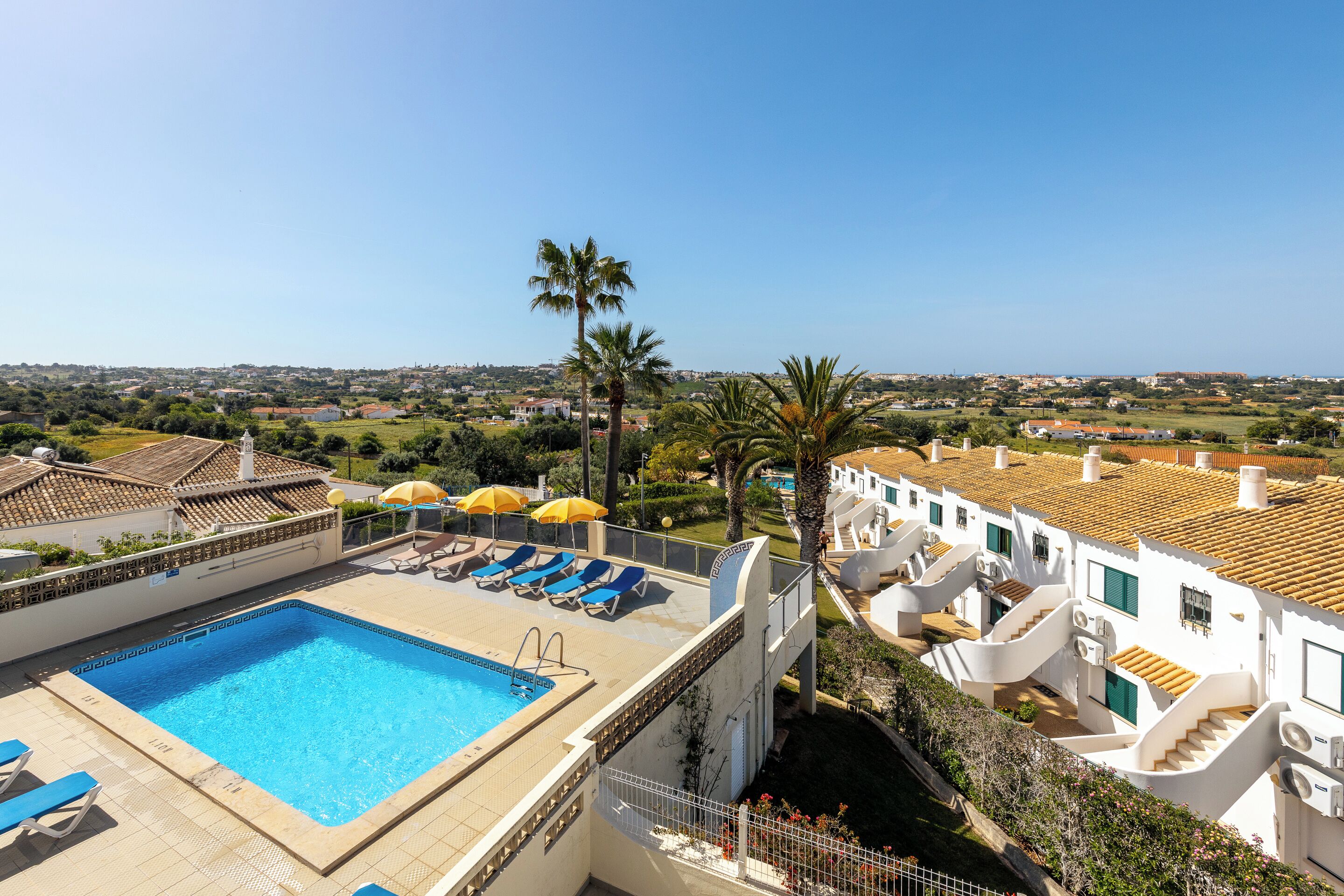 A rooftop pool with blue water, surrounded by white buildings with red-tiled roofs, and a clear view of the distant landscape.