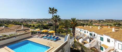 A rooftop pool with blue water, surrounded by white buildings with red-tiled roofs, and a clear view of the distant landscape.