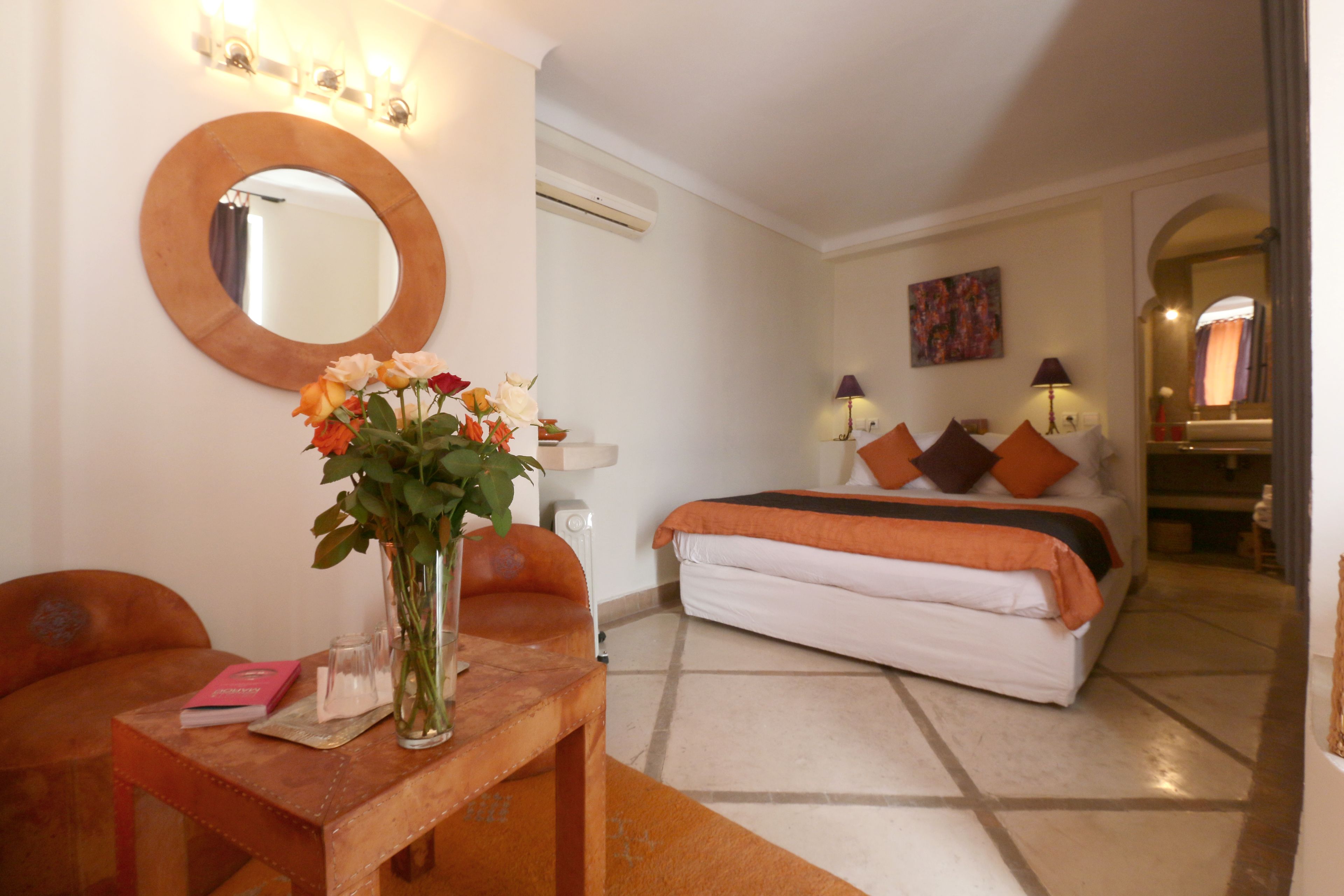 Un dormitorio con una cama, una mesita de noche con un florero, un espejo redondo y una lámpara.