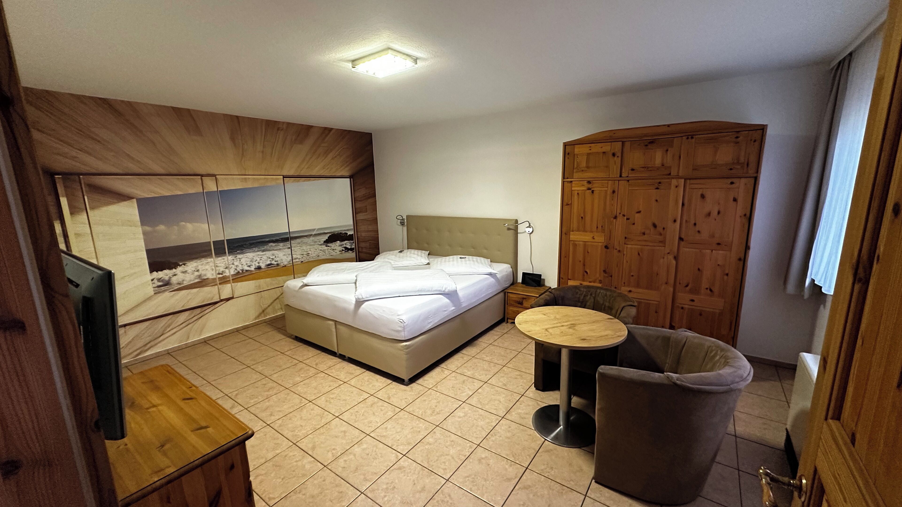 Een slaapkamer met een bed, een houten kledingkast, een klein rond tafeltje, een stoel en een televisie.