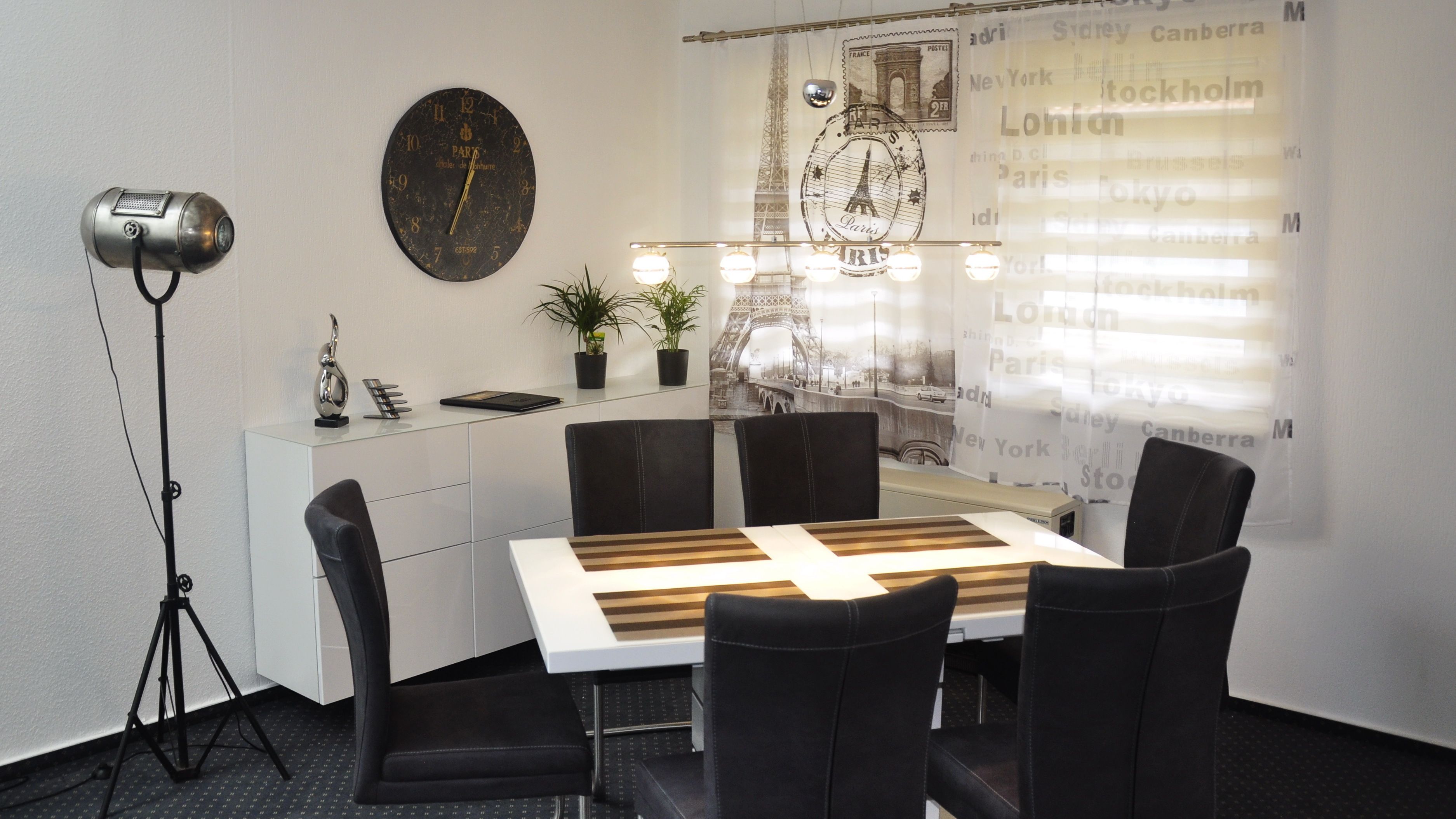 Un comedor con una mesa, sillas, un reloj, una planta en maceta y una pared con nombres de ciudades.
