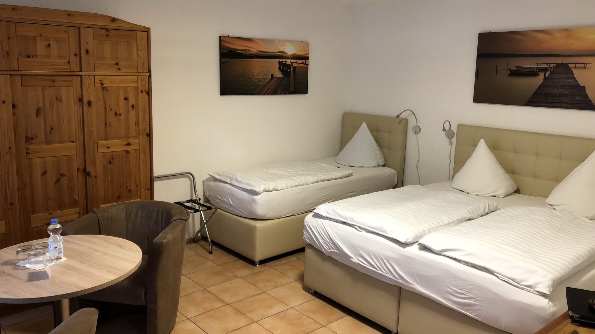 Une chambre d’hôtel avec deux lits, une armoire en bois, une petite table ronde avec une bouteille d’eau, et deux tableaux accrochés au mur représentant un coucher de soleil sur l’eau.