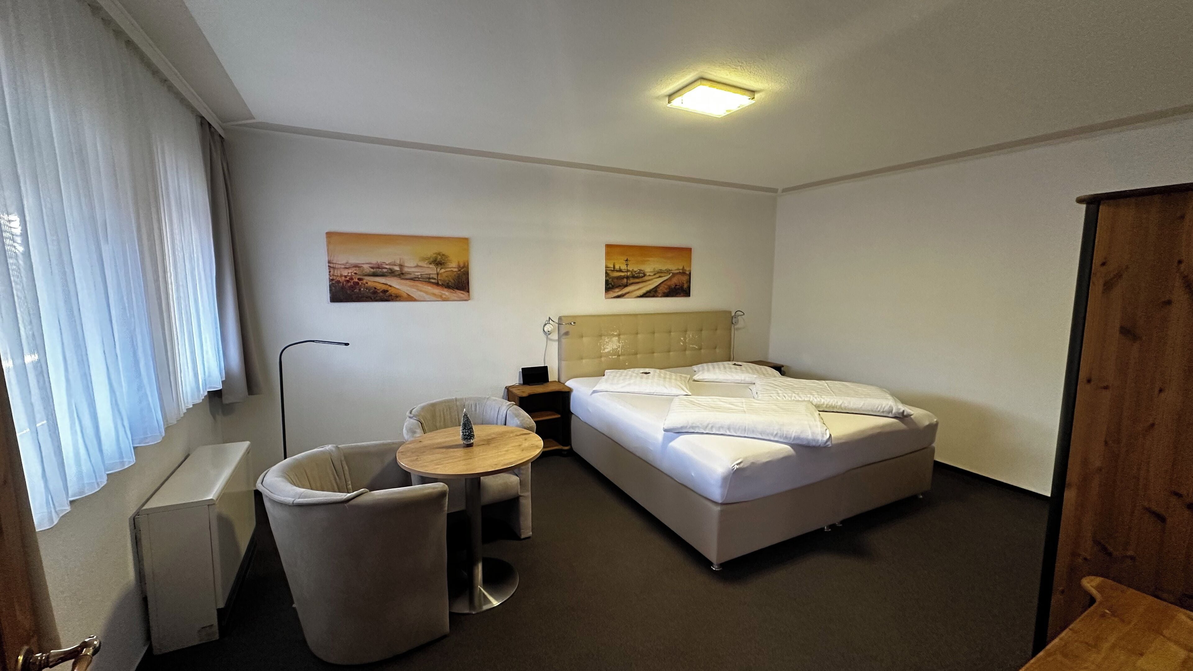 Quarto de hotel com cama, escrivaninha, cadeira, mesinha e duas pinturas na parede.