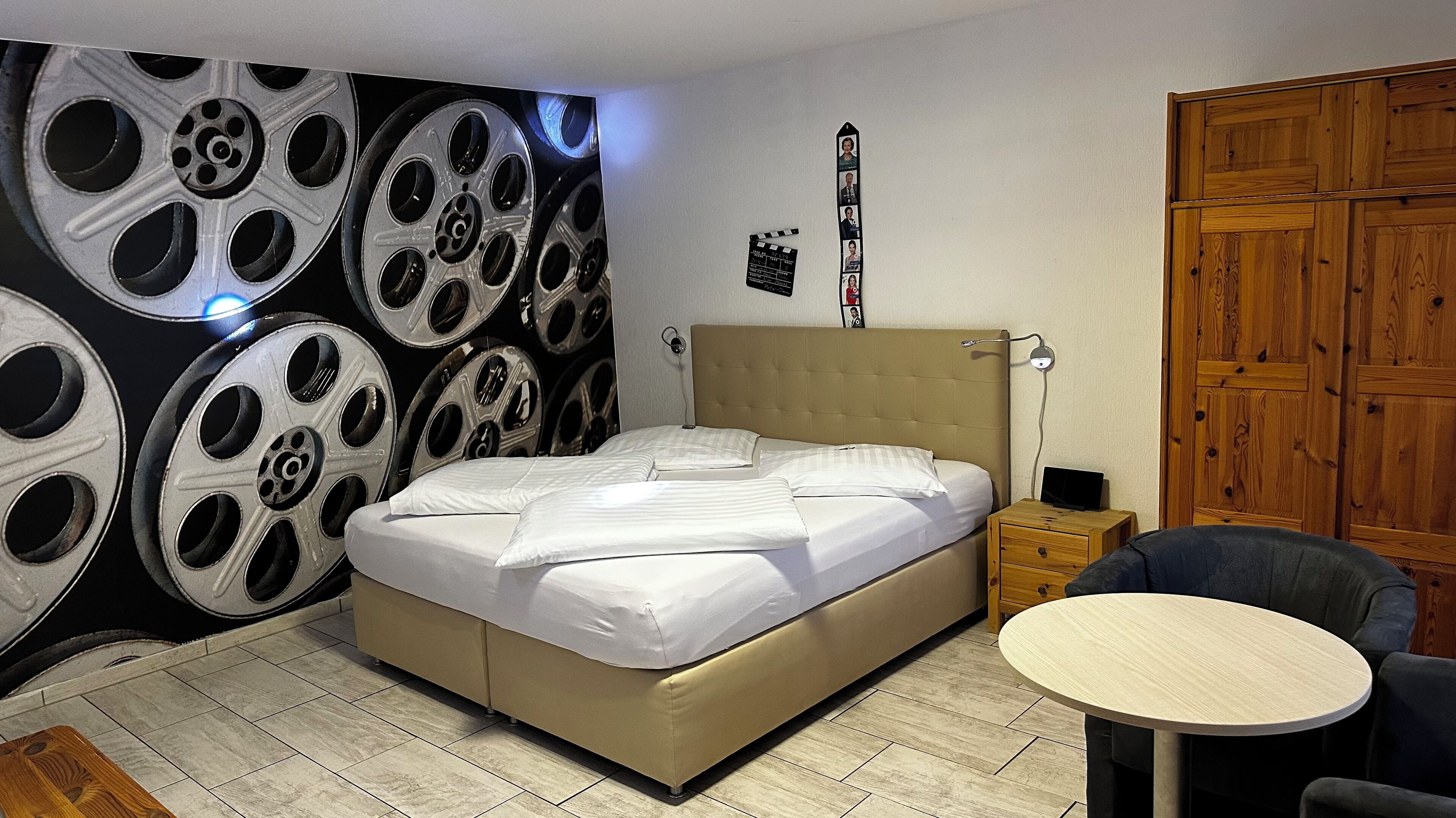 Ein Schlafzimmer mit einem Bett, einem Nachttisch, einem Sessel, einem runden Tisch, einer Wand mit Filmrollen-Design und einem Holzschrank.