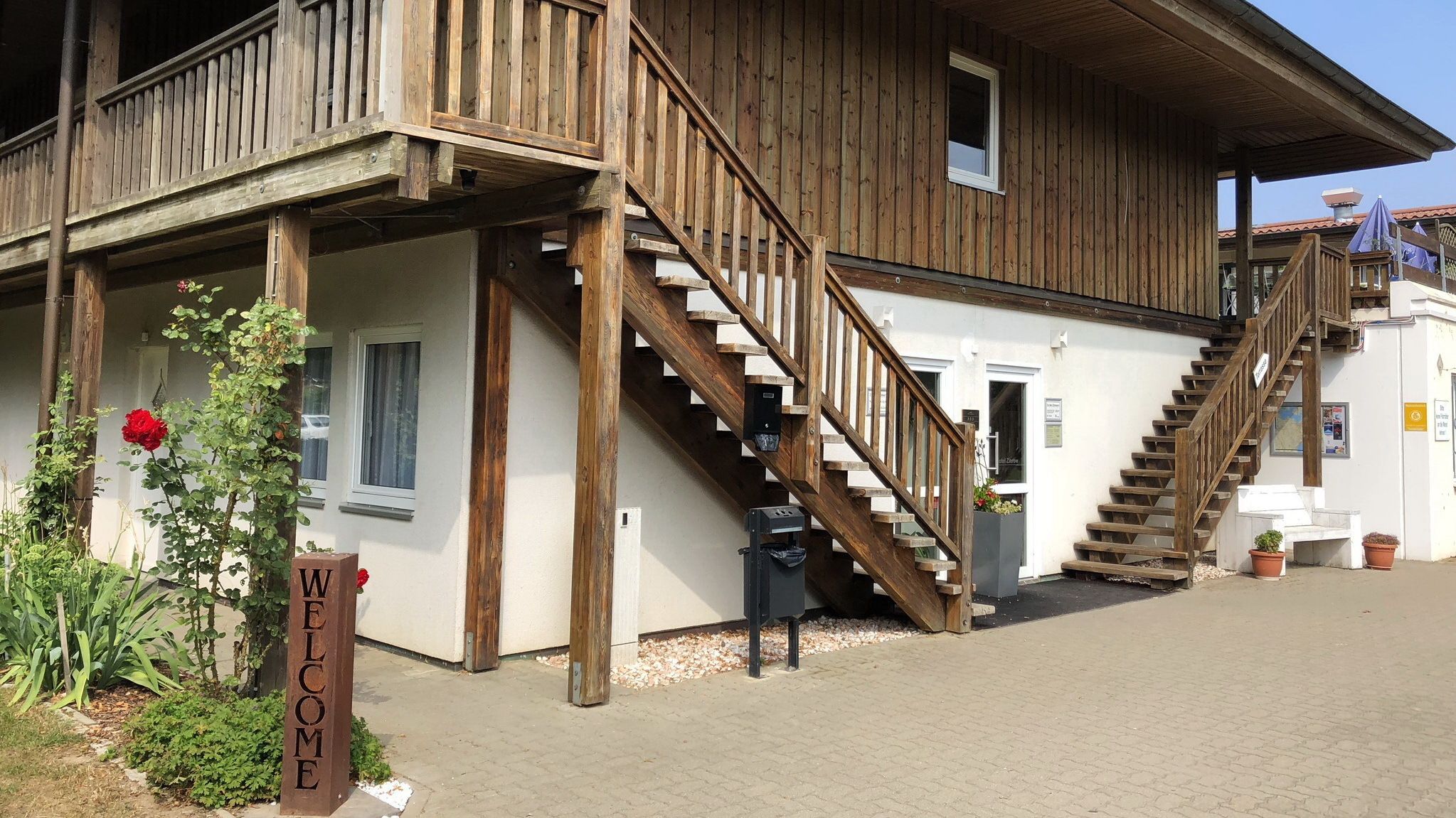 Ein hölzernes Gebäude mit freundlicher Beschilderung, einer Treppe und einem Garten mit einer roten Rose.