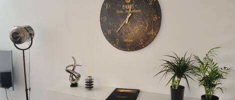 A wall clock with a Paris design, a desk with a book, a camera, and two potted plants.