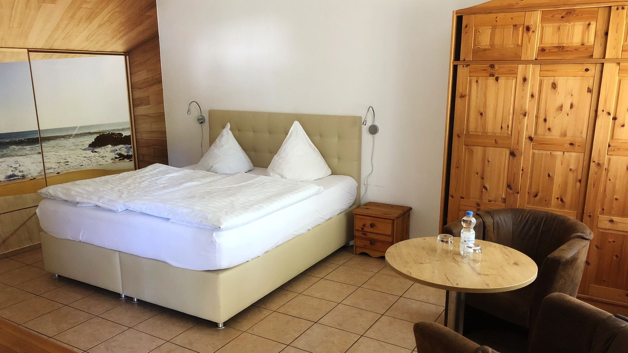 A bedroom with a bed, a nightstand, a round table with a chair, and a wooden wardrobe.