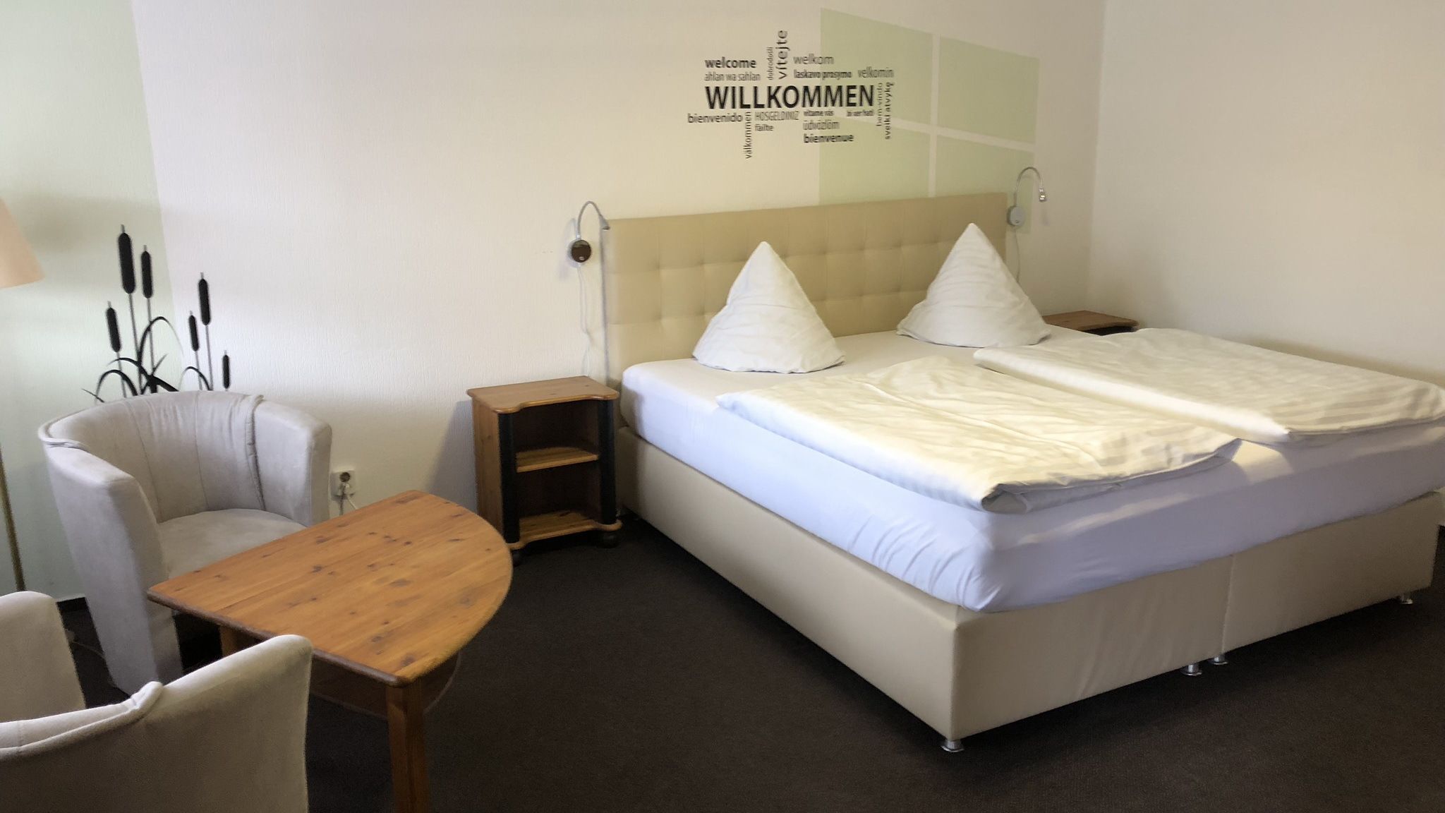 Una habitación de hotel con una cama, una silla, una mesita de madera y una pared con carteles de bienvenida en varios idiomas.