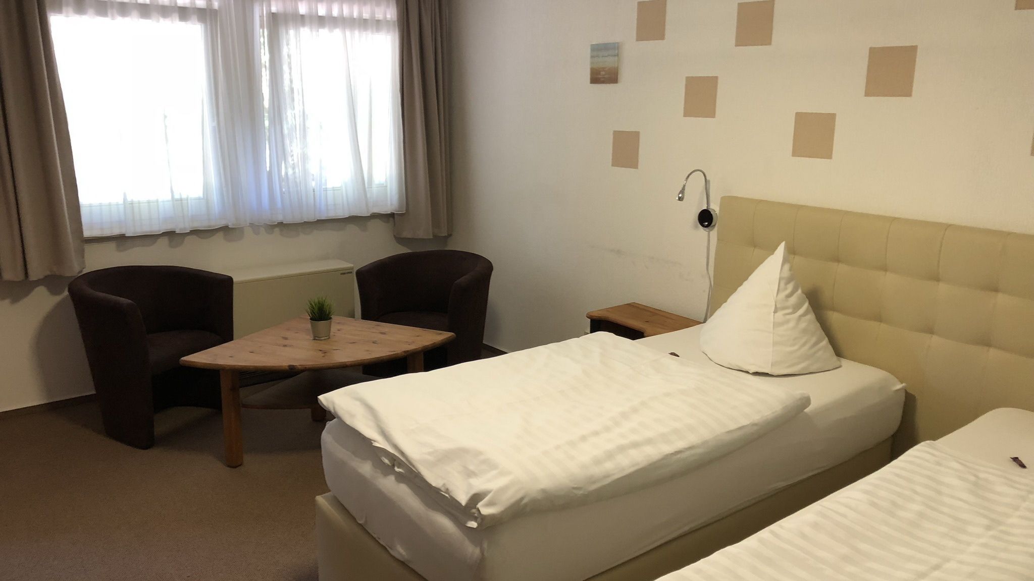 Quarto de hotel com duas camas de solteiro, uma mesinha com uma planta, duas cadeiras, um criado-mudo e uma luminária na parede.