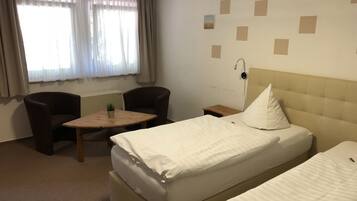 Quarto de hotel com duas camas de solteiro, uma mesinha com uma planta, duas cadeiras, um criado-mudo e uma luminária na parede.