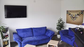Ein Wohnzimmer mit einem blauen Sofa, einem kleinen Holztisch, einem Topfpflanzen und einem flachbildschirmigen Fernseher, der an der Wand befestigt ist.