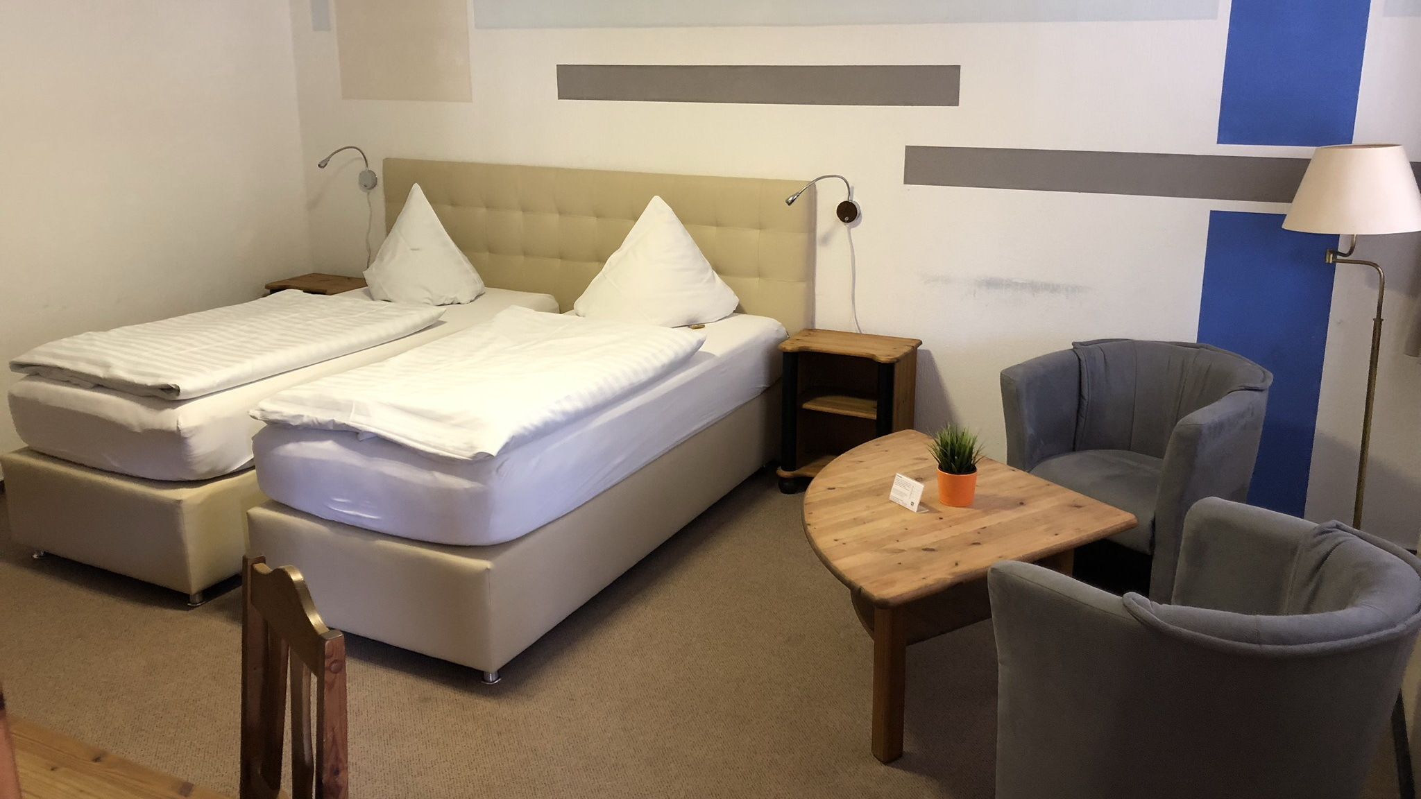 Ein Hotelzimmer mit einem Doppelbett, zwei Nachttischen, einem kleinen Holztisch mit einer Pflanze, einem grauen Sessel und einer Lampe.