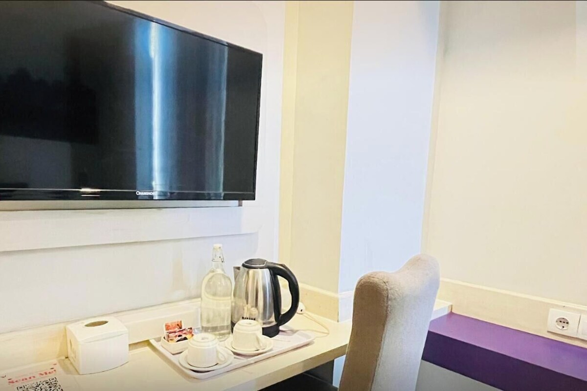A hotel room with a flat-screen TV mounted on the wall, a small table with a kettle, a bottle, and a cup, and a chair with a cushion. The room has a simple layout with a purple accent wall.