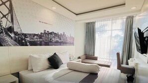 Une chambre d’hôtel avec un grand lit, une chaise, un bureau et une télévision. La chambre est décorée d’une fresque représentant un paysage urbain sur un des murs, et elle possède une fenêtre avec des rideaux.