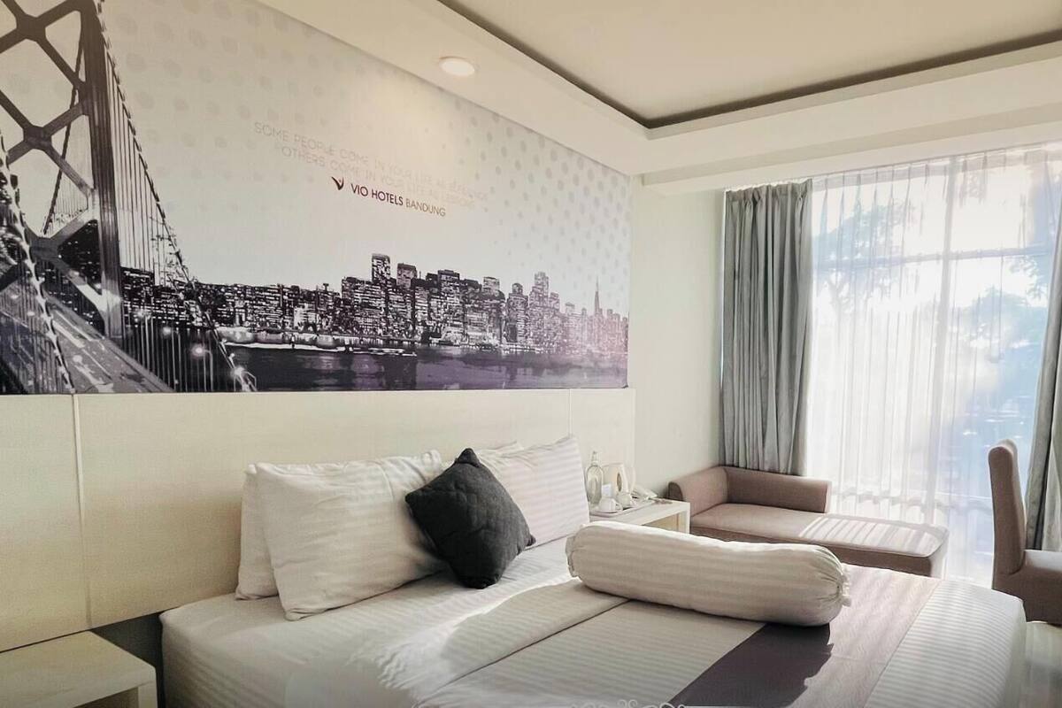 A hotel room with a large bed, a chair, a desk, and a TV. The room has a cityscape mural on the wall and a window with curtains.