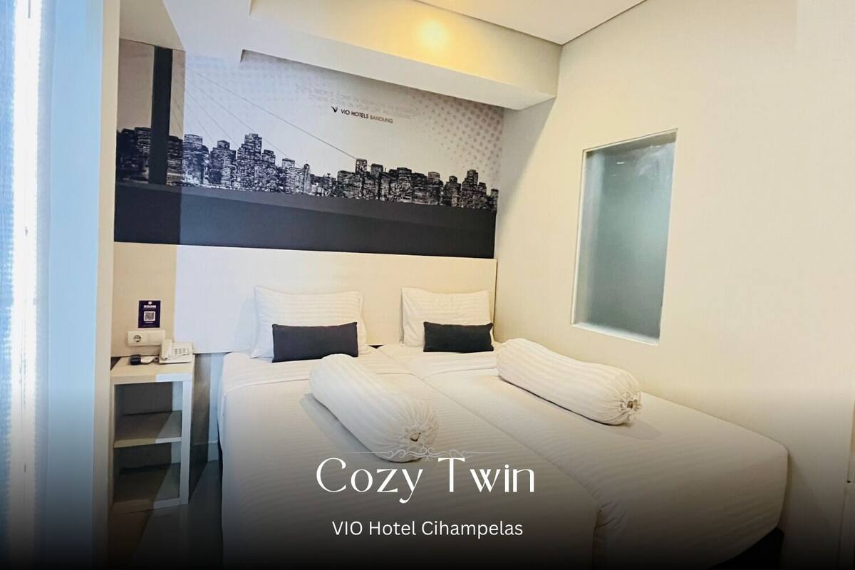 A hotel room with a cityscape mural on the wall, a bed with two pillows, and a bedside table with a phone.