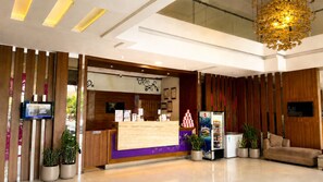 A reception area with a wooden reception desk, a seating area with a sofa, potted plants, and a ceiling with recessed lighting. - Vio Cihampelas (Bandung)