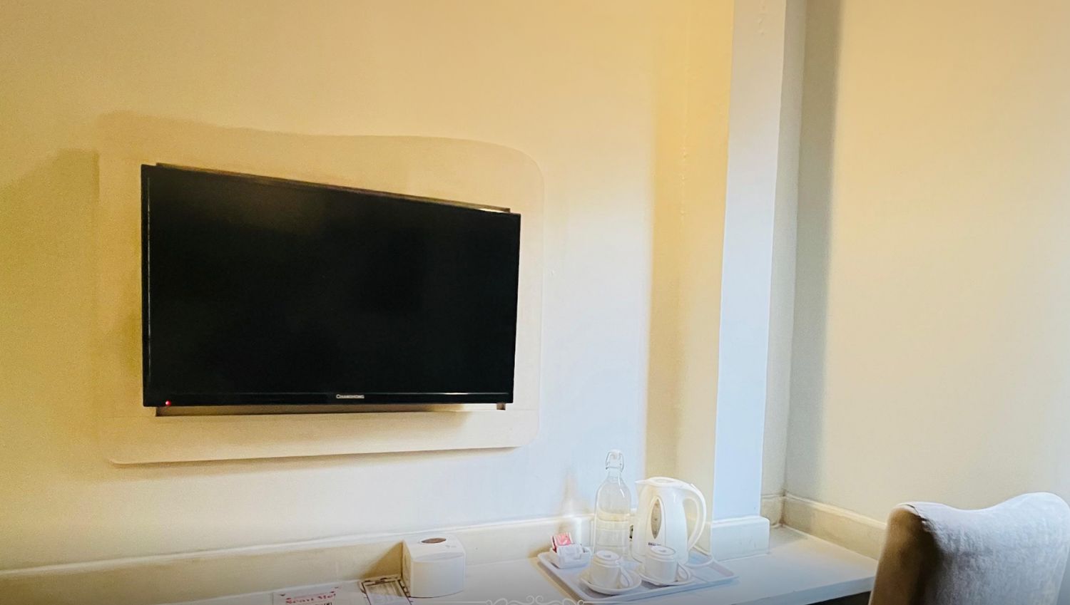 A hotel room with a mounted television, a small table with a tray of tea and cups, and a chair. The room is labeled "Comfort Queen" at the VIO Hotel Cihampelas.