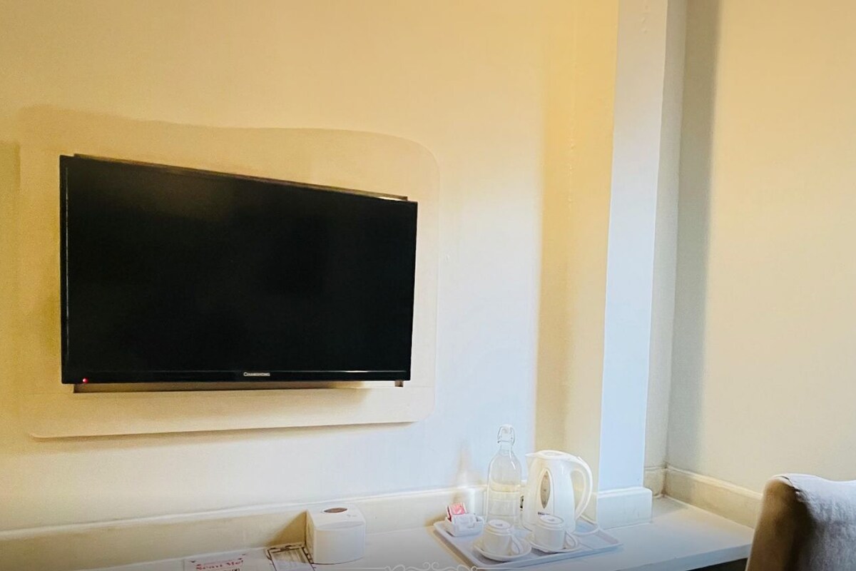 A hotel room with a mounted television, a small table with a tray of tea and cups, and a chair. The room is labeled "Comfort Queen" at the VIO Hotel Cihampelas.