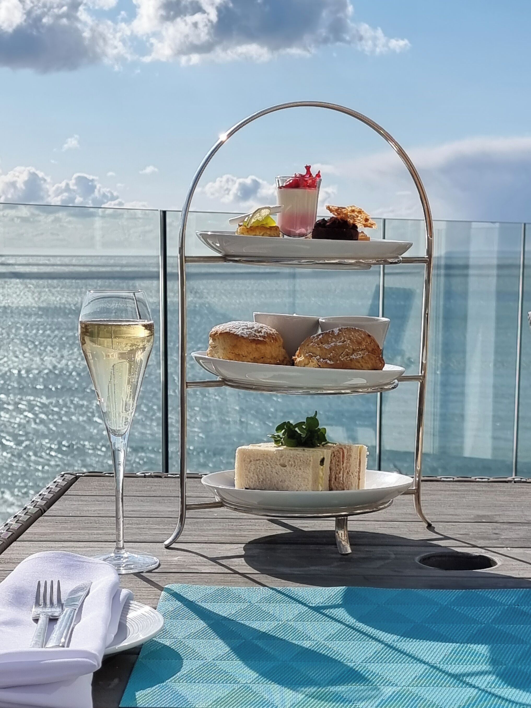 Ett dessertfat i tre våningar med olika sorters bakverk och ett glas vitt vin, placerat på en balkong med utsikt över havet.