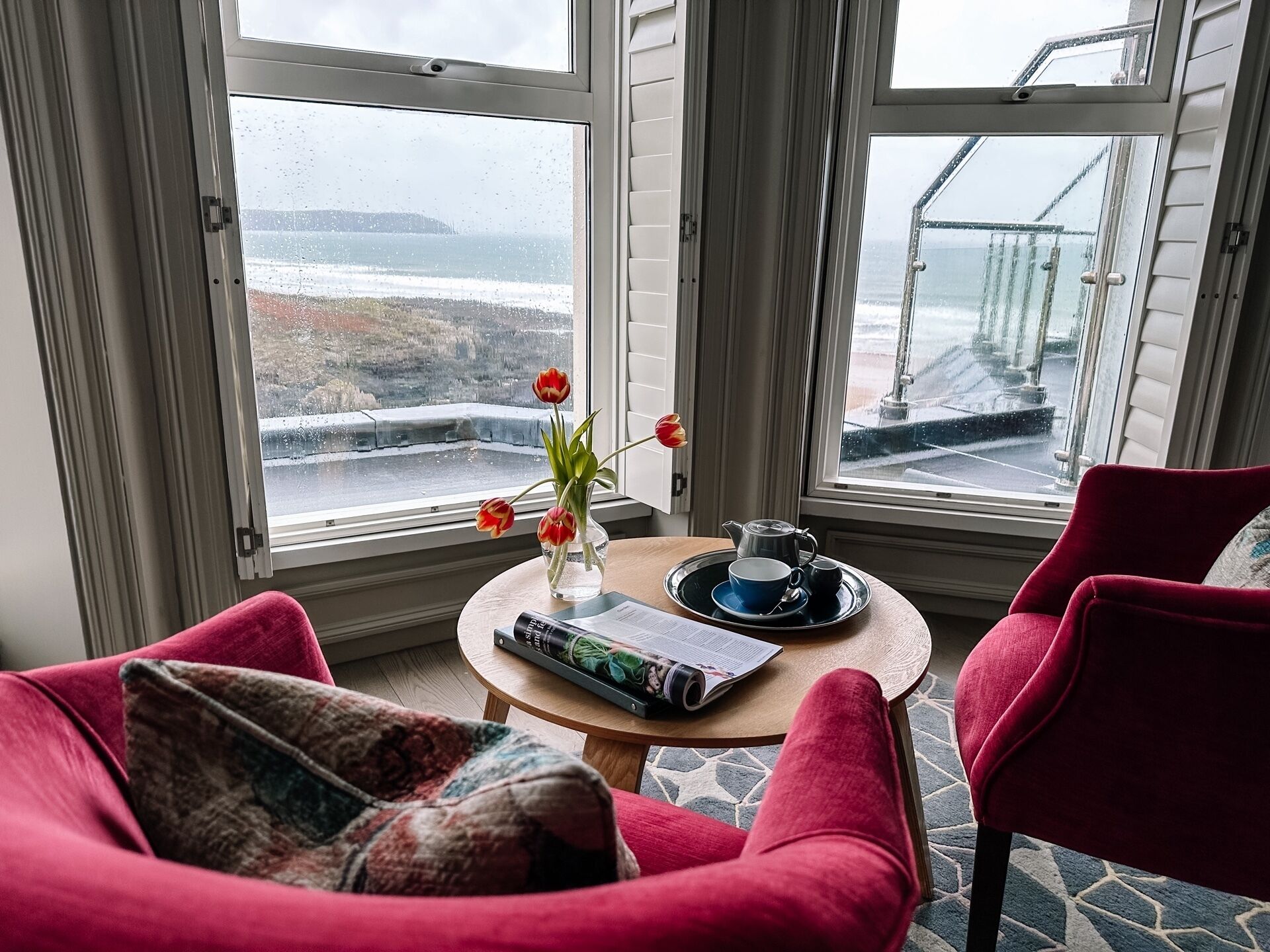 A cozy room with a view of the sea, featuring a red chair, a small table with a book and a vase of flowers.