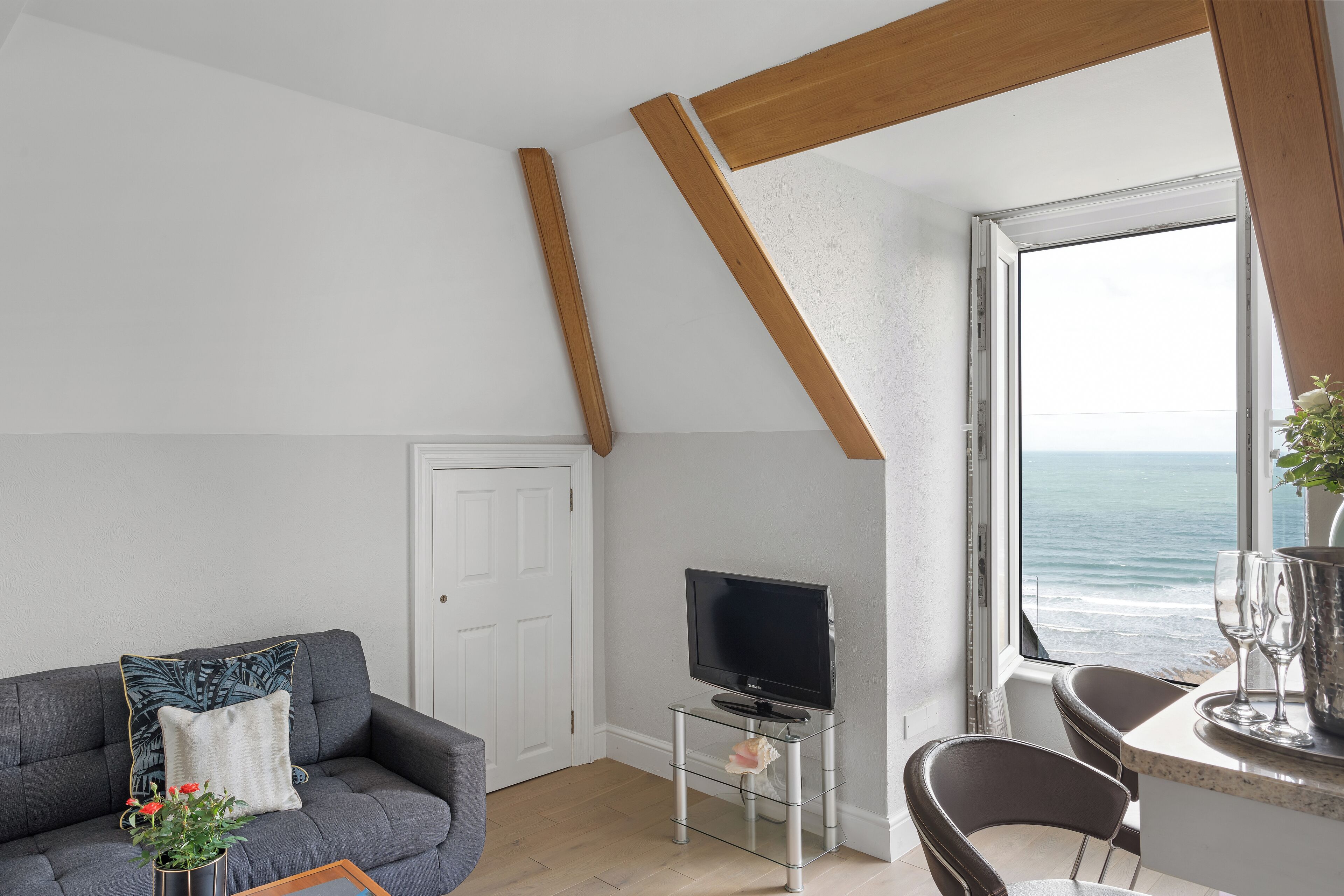 Ein modernes Wohnzimmer mit grauem Sofa, einem Fernseher auf einem Glastisch, einem Essbereich mit Tisch und Stühlen sowie Meerblick durch ein großes Fenster.