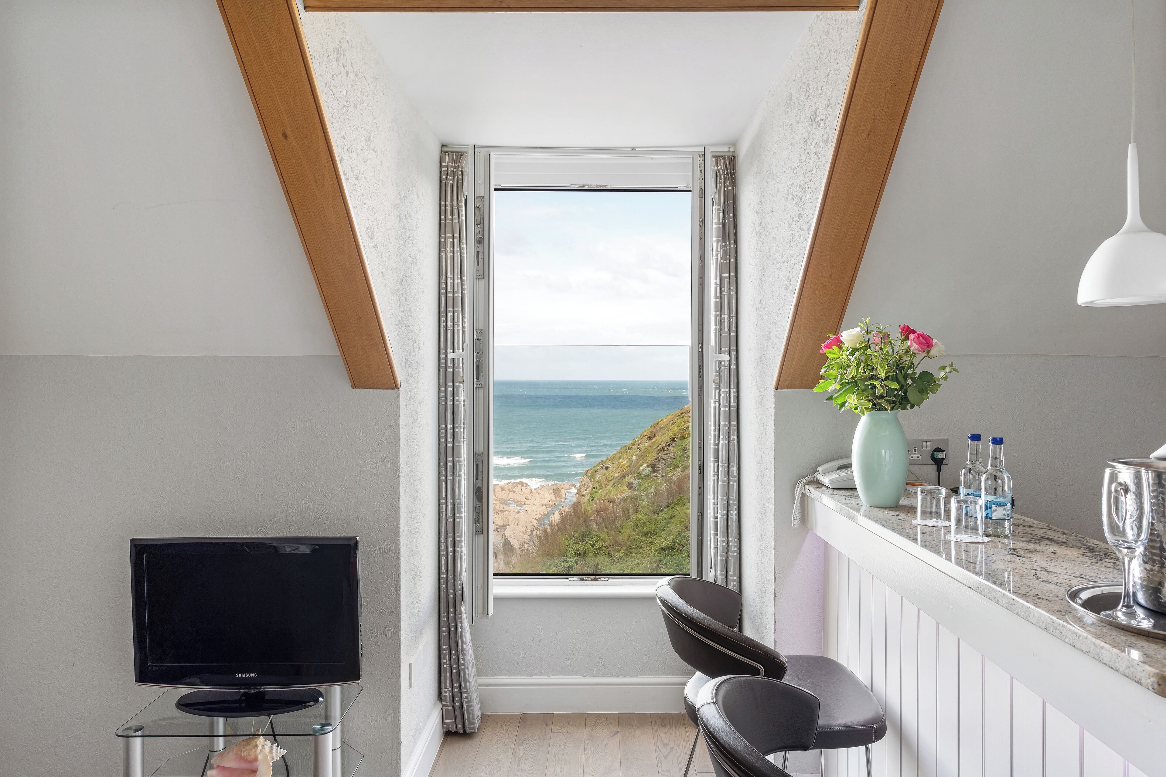 Une pièce avec un téléviseur posé sur un meuble en verre, un coin bar agrémenté d’un vase de fleurs et une vue sur la mer par la fenêtre.