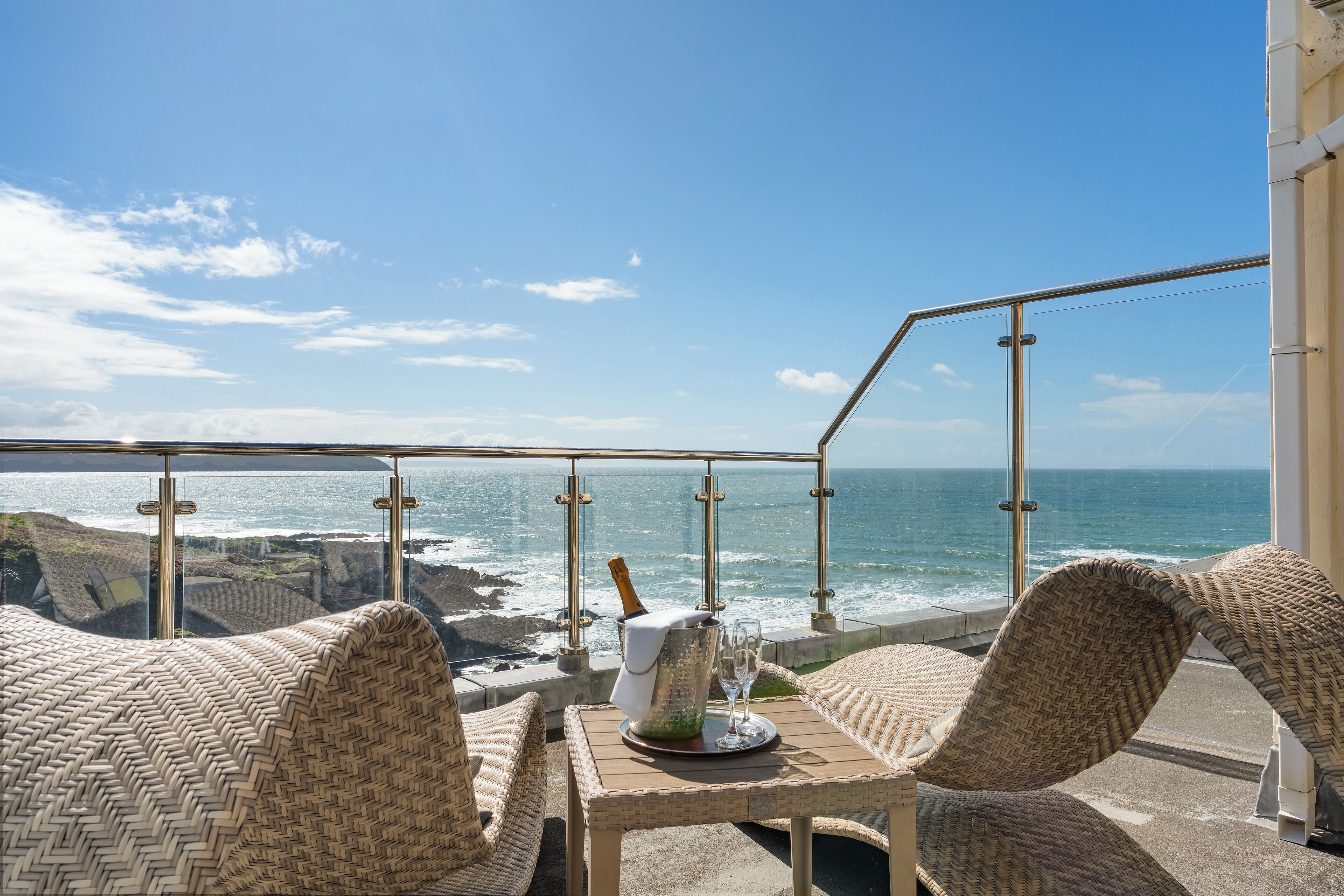 A balcony with wicker furniture, a table with a bottle and glasses, and a view of the ocean.