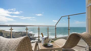 A balcony with wicker furniture, a table with a bottle and glasses, and a view of the ocean.
