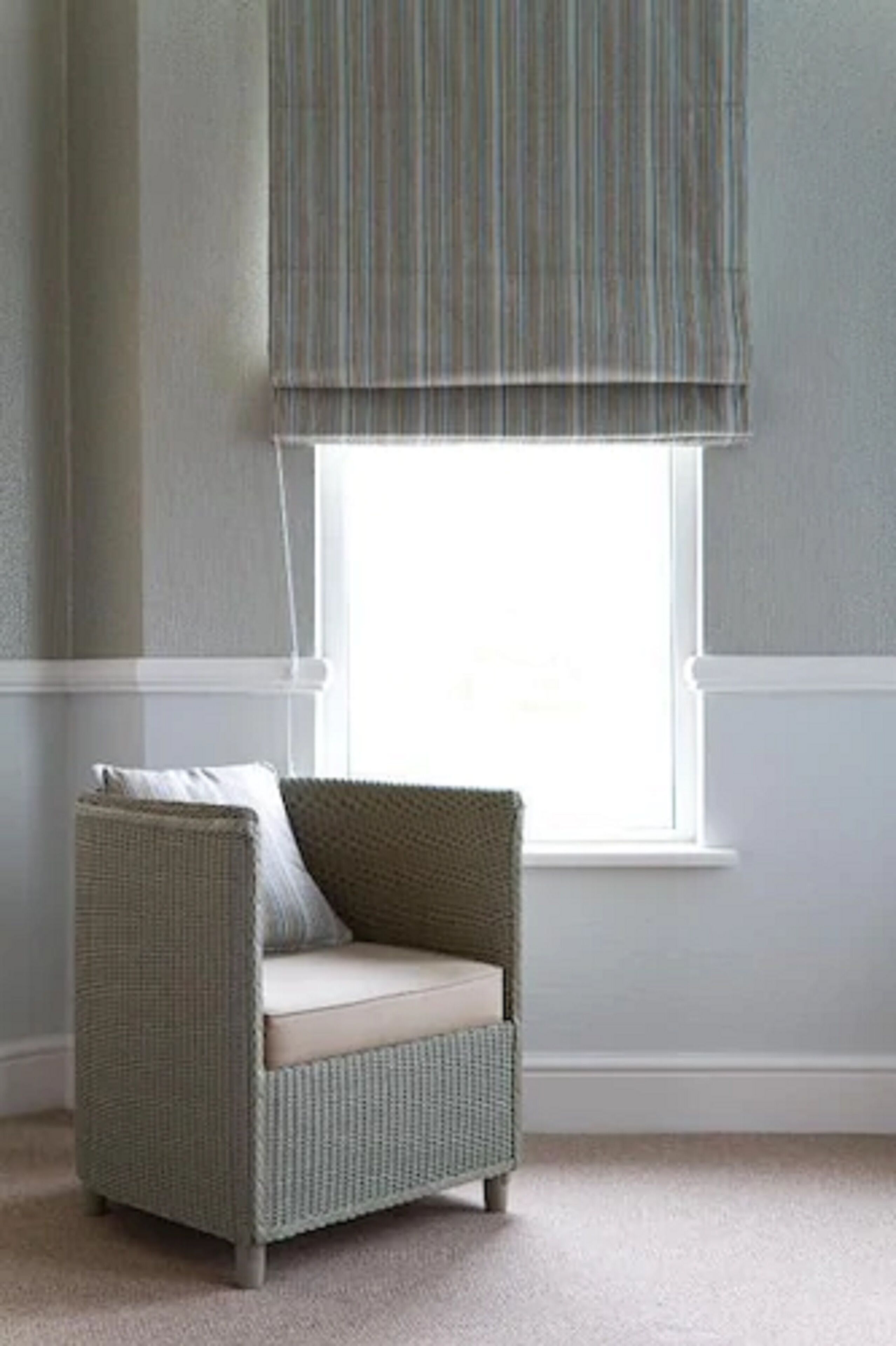 Un sillón de mimbre con un cojín, ubicado frente a una ventana con persiana a rayas.