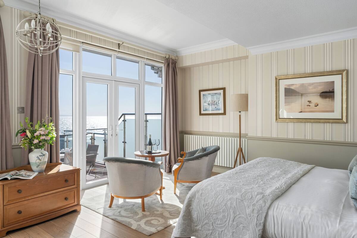 Ein Hotelzimmer mit einem großen Bett, zwei Sesseln, einer Holzkommode, einem Balkon mit Tisch und Stühlen sowie Blick auf das Meer.