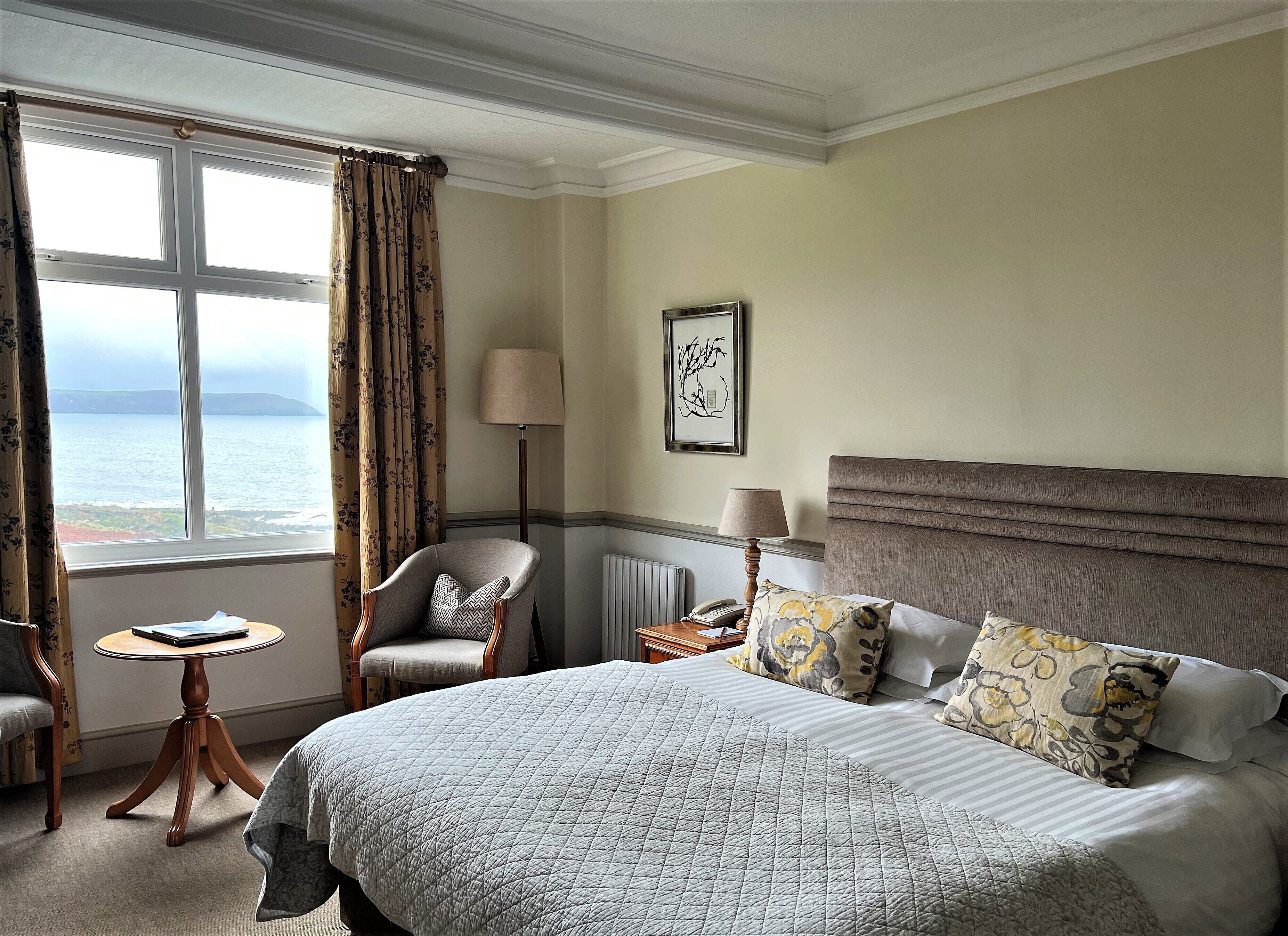 Habitación de hotel con una cama grande, una silla, una mesita, una lámpara y vistas al mar.