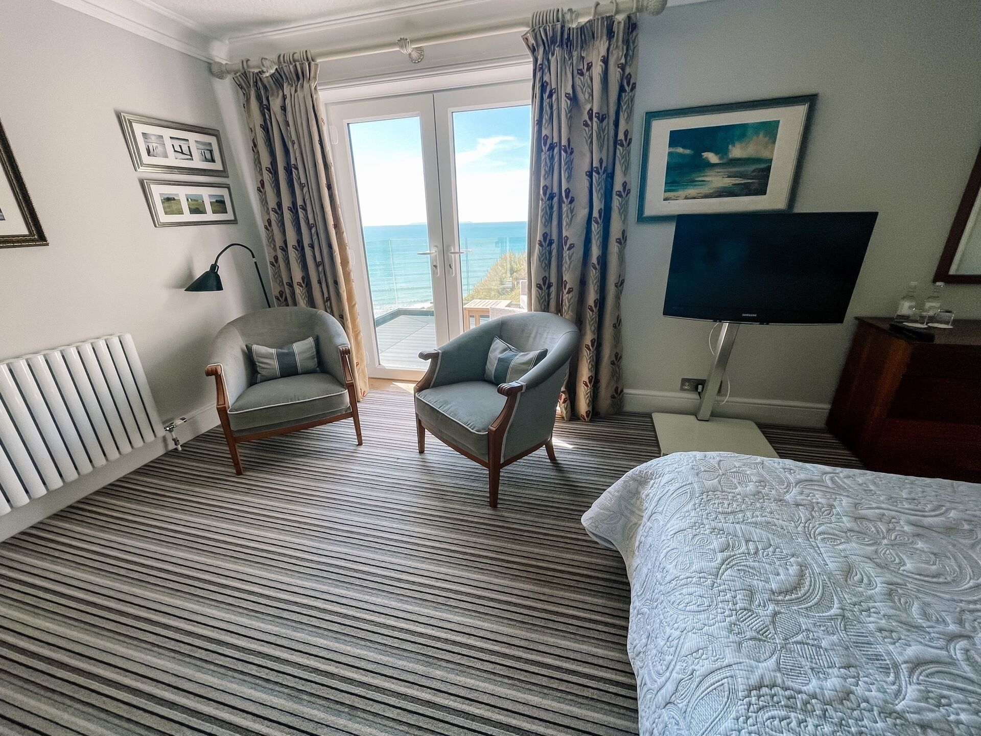 A hotel room with a bed, two armchairs, a TV, a radiator, and a view of the sea through the window.