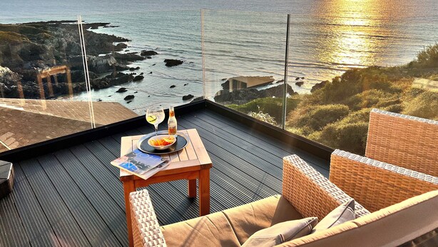 Una terraza con una mesa dispuesta para dos, con vista a un paisaje costero de costas rocosas y una vista al atardecer.