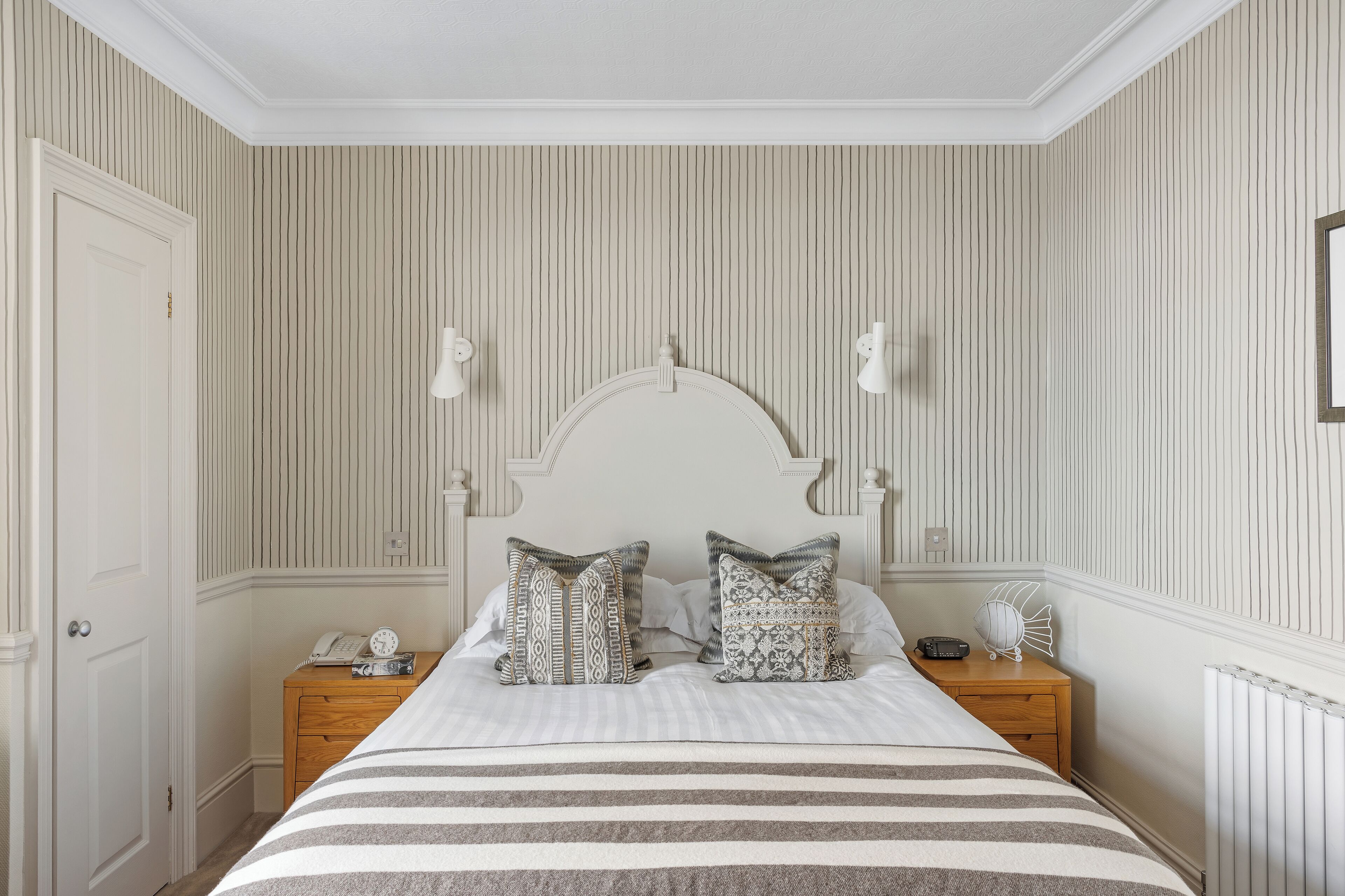 A neatly arranged bedroom with a bed, two bedside tables, a nightstand, and a wall-mounted light fixture.