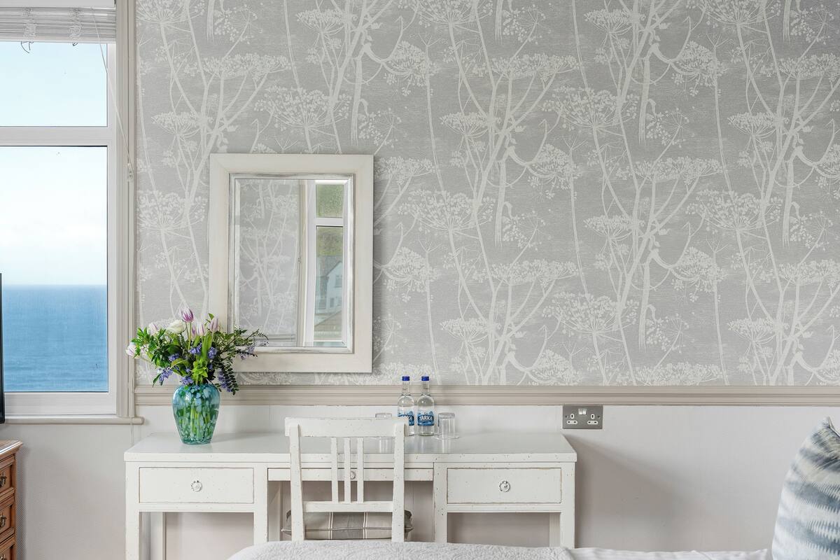 Ein Zimmer mit weißem Schreibtisch, einem Spiegel, einer Vase mit Blumen und Meerblick durch ein Fenster.