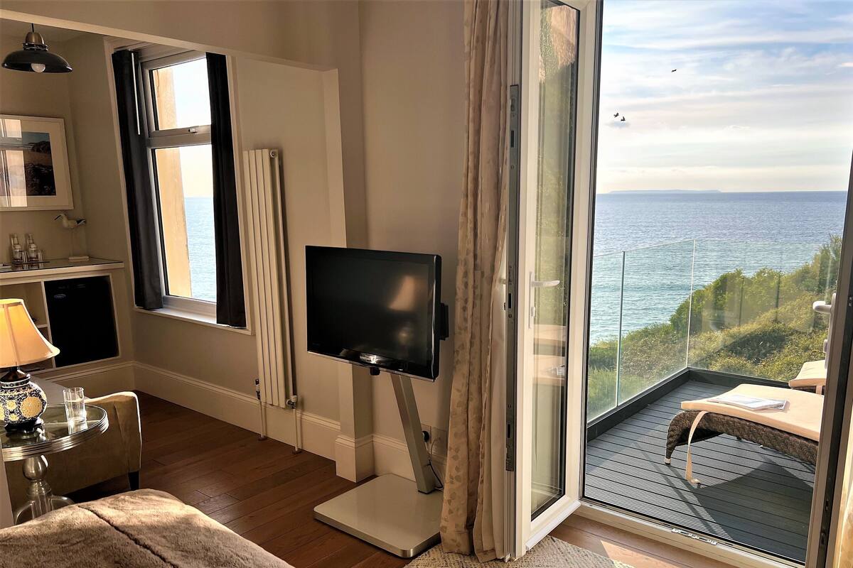 Ein Zimmer mit Fernseher, einem kleinen Tisch mit Lampe und Meerblick durch eine Glastür.