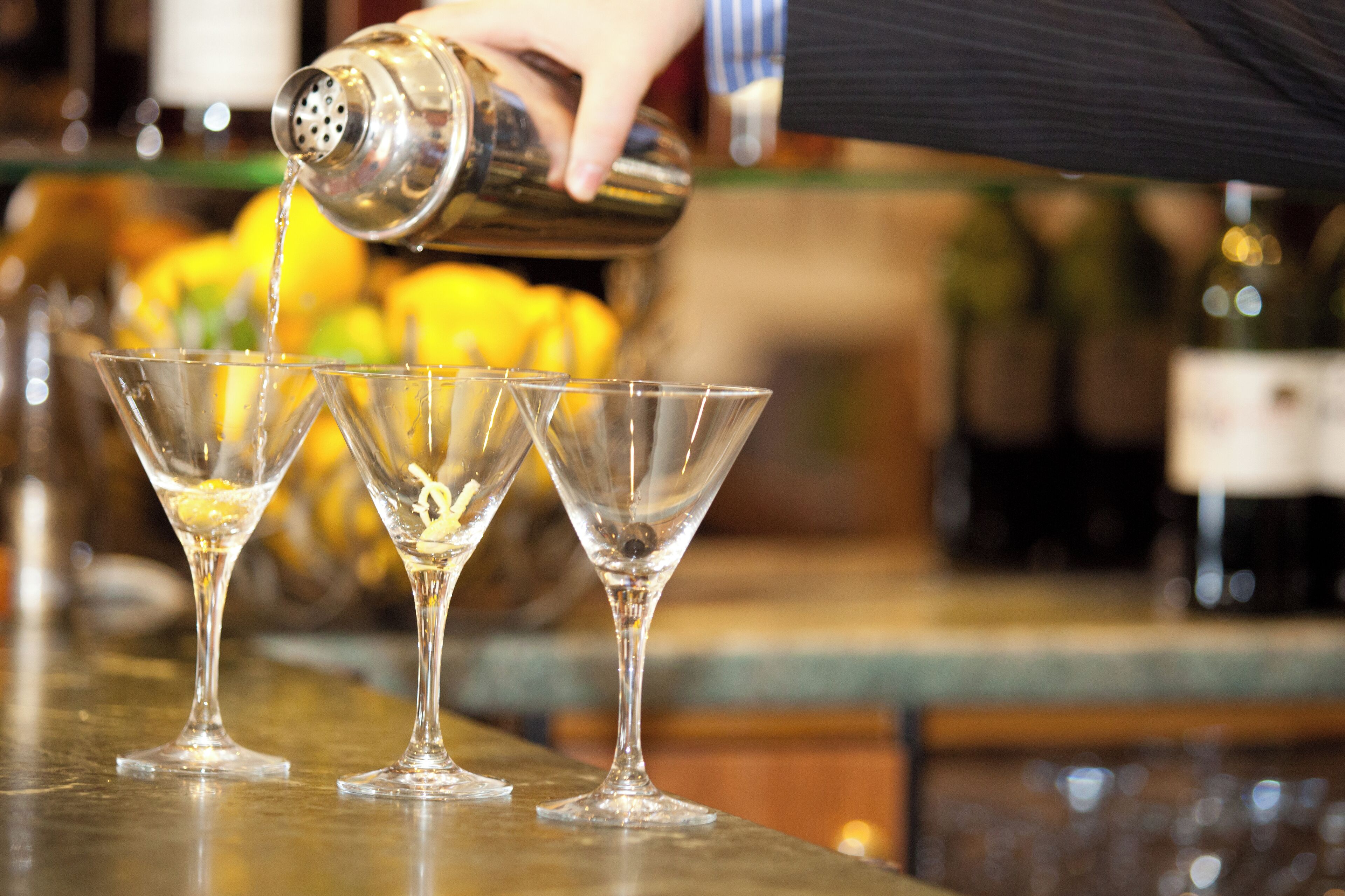A person pouring a drink into three martini glasses on a bar.