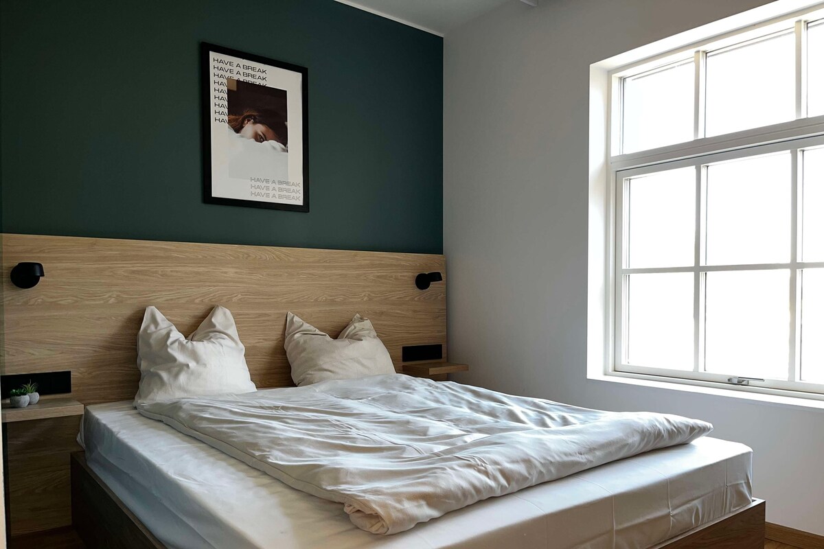 A neatly made bed with a wooden headboard, a bedside table with a lamp, and a window with a view of the outside.