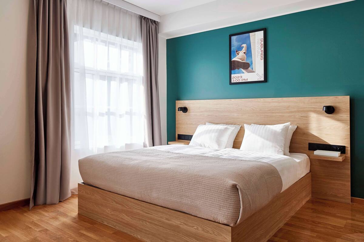 Moderni hotellihuone, jossa on puinen sänky, turkoosinsävyinen korosteseinä ja ikkuna, jossa on läpikuultavat verhot.