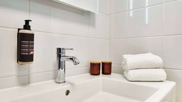Un baño con lavamanos blanco, grifo cromado, dispensador de jabón, dos tazas de madera y dos toallas blancas dobladas.