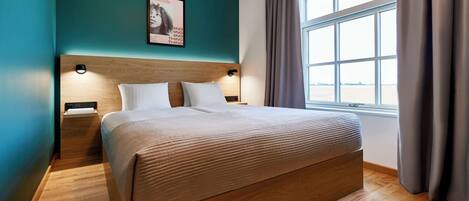 Un lit bien fait dans une chambre aux murs de couleur turquoise, avec une tête de lit en bois et une fenêtre agrémentée de rideaux.