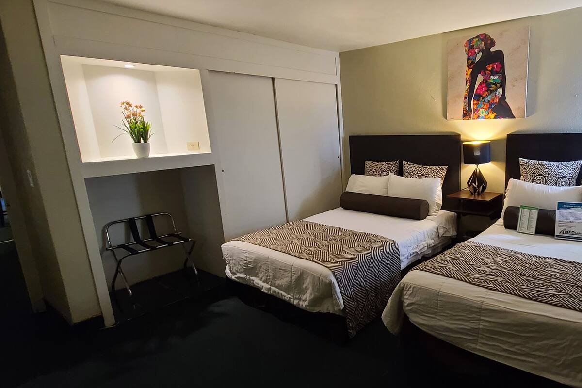 A hotel room with two beds, a small desk, a nightstand with a lamp, a wall-mounted shelf with a plant, and a painting above the beds.